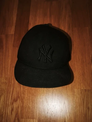 Svart New Era NY keps - Säljer en svart keps från New Era med broderad NY-logga framtill. Klassisk modell med böjd skärm och diskret New Era-logo på sidan. Kepsen är helsvart och har en stilren look som passar till det mesta. Materialet känns slitstarkt och kepsen har ventilerande hål upptill.