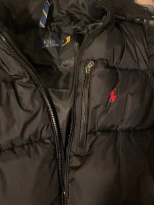 Svart pufferjacka från Polo Ralph Lauren - Snygg svart pufferjacka från Polo Ralph Lauren med röd broderad logga på bröstet. Jackan har dragkedja framtill, flera fickor med dragkedja och hög krage. Perfekt för kalla dagar och riktigt skön att bära. Klassisk design som alltid funkar. Pris kan diskuteras 