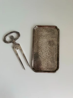 Antikt uppläggningsset i nysilver - Säljer ett antikt uppläggningsset i nysilver med dekorativt handtag och rektangulärt fat. Gaffeln har vackra detaljer och stämpel, perfekt för servering eller som samlarobjekt. Båda delarna har klassisk vintagekänsla och passar till dukning med retrostil.