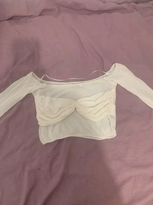 Vit croppad mesh topp med lång ärm - Trendig vit croppad topp i mesh med långärmad design och twistad detalj framtill. Toppen har en offshoulder-look och är perfekt för att styla med höga jeans eller kjol. Tunn och lätt, passar bra till varmare dagar.