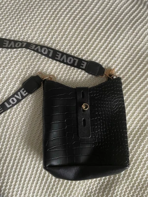 Svart axelväska med croco-mönster - Cool svart axelväska i croco-mönstrad konstläder med bred axelrem där det står LOVE i vitt. Väskan har guldfärgade detaljer och en praktisk form som passar till många outfits. Perfekt för dig som gillar trendiga accessoarer.