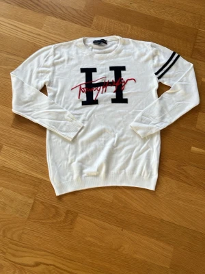Vit stickad tröja Tommy Hilfiger - Vit stickad tröja från Tommy Hilfiger med stort svart H och röd logga framtill. Tröjan har långa ärmar och två svarta ränder på ena ärmen. Ribbstickade muddar vid ärmslut och nederkant. Perfekt för en clean och sportig look. Storleken är 164-170 men passar snarare 153-160