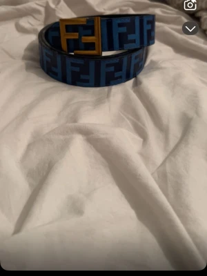 Blått Fendi bälte med FF-logga - Säljer ett blått bälte från Fendi med ikoniskt FF-mönster i mörkblått och en stor guldfärgad FF-spänne framtill. Bältet är gjort i läder och har en modern, trendig look som sticker ut. Perfekt accessoar för att lyfta vilken outfit som helst.