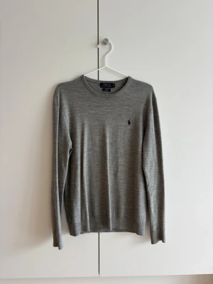 Polo Ralph Lauren Merino Pullover - Snygg grå stickad tröja från Polo Ralph Lauren med rund halsringning och diskret mörkblå logga broderad på bröstet. Tröjan har långa ärmar och är tillverkad i mjukt, tunt material som passar perfekt till lager på lager.