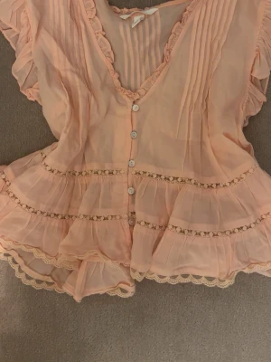 Koralfärgad volangblus med spets - Koralfärgad blus med volanger, spetsdetaljer och knappar framtill. Blusen har v-ringning, korta ärmar och är gjord i ett tunt, transparent tyg som är luftigt. Perfekt för sommaren och finare tillfällen. 