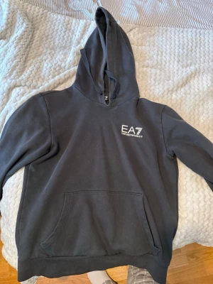 Svart hoodie från EA7 Emporio Armani - Snygg svart hoodie från EA7 Emporio Armani med vit logga på bröstet. Klassisk känguruficka framtill och justerbar huva med snören. Tillverkad i mjukt bomullsmaterial som är skönt att ha på sig. Perfekt för en avslappnad och trendig look.