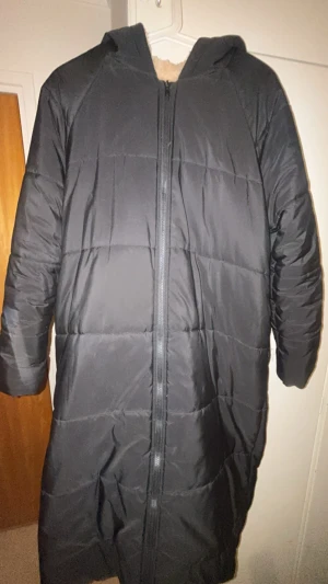 Svart lång dunjacka med huva - Säljer en svart lång dunjacka med huva och dragkedja framtill. Jackan har quiltad design och är fodrad med mjukt material i huvan. Perfekt för kalla dagar och riktigt skön att bära. Passar dig som vill ha en stilren och varm jacka i vinter. Använd några fåtal gånger.
