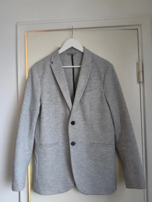 Ljusgrå kavaj från Zara - Stilren ljusgrå kavaj från Zara med två knappar framtill och klassisk krage. Kavajen har två framfickor och en bröstficka, samt mörkblått innerfoder. Perfekt för en trendig och clean look.