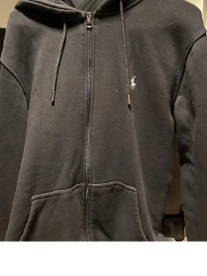 Svart hoodie med dragkedja från Polo - Svart hoodie med dragkedja från Polo Ralph Lauren. Klassisk design med huva och dragsnören, två fickor framtill och den ikoniska vita Polo-loggan broderad på bröstet. Perfekt för en avslappnad och stilren look.