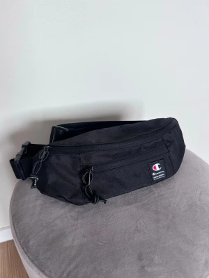 Svart streetstyle y2k crossbody bag från Champion - Svart crossbody väska från Champion med många olika fack och väldigt snygg design. Bär den runt midjan, tvärs över bröstet eller över axeln. Sälj pga att den inte används. Nyskick!