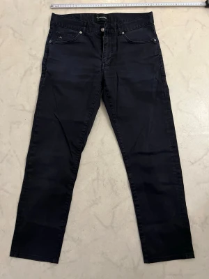 Svarta jeans från J.Lindeberg Jay - strl.31/32 - Säljer ett par svarta jeans från J.Lindeberg med klassisk femficksmodell och raka ben. Jeansen har silverfärgade nitar och knapp, samt diskret J.Lindberg-logga på fickan. Materialet är mjukt och stretchigt för extra komfort.