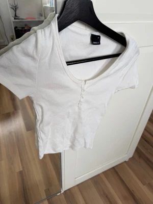 Vit ribbad topp med knappar - Säljer en vit ribbad topp från Monki med korta ärmar och v-ringning. Toppen har små knappar framtill och är croppad i modellen. Perfekt till jeans eller kjol för en enkel och snygg look.