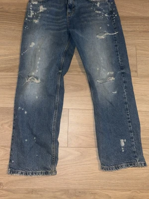 Blå baggy jeans med slitningar - Säljer ett par blå baggy jeans med slitningar och färgstänk för en cool, avslappnad look. Jeansen har raka ben, klassisk femficksmodell och tydliga slitdetaljer på framsidan. Perfekta för dig som gillar streetwear och vintage vibe.