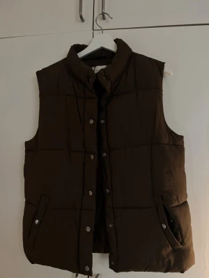 Mörkbrun dunväst med hög krage - Snygg mörkbrun dunväst med hög krage och tryckknappar framtill. 