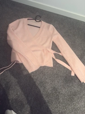 Ljusrosa omlottstickad tröja - Supersöt ljusrosa stickad omlott-tröja med långa ärmar och knytband i midjan. Tröjan har v-ringning och ribbade muddar vid ärmslut och nederkant. Perfekt för lager-på-lager och en mjuk känsla.