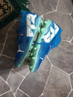 Blåa Puma fotbollsskor med dobbar -  Säljer ett par blåa fotbollsskor från Puma med vita detaljer och logga på sidan. Skorna har snörning, högt skaft och ljusgröna dobbar för bra grepp på planen. Tillverkade i syntetmaterial. Det går tyvärr inte att se storleken så bra men det är 39,5. Använd fåtal gånger fast fortfarande i mycket gott skick. Om du vill ha en av bollarna pumpade kostar det 10kr extra för jag behöver åka någonstans för och göra det! PRIS KAN DISKUTERAS! Vad ingår: en fotboll om du skulle velat ha det sen skorna.