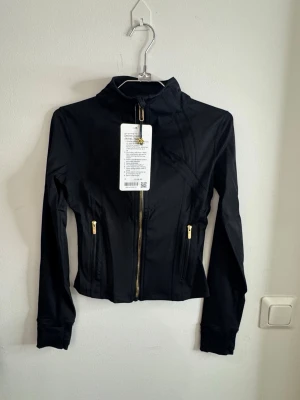 Svart träningsjacka från Lululemon storlek 4 - Snygg svart träningsjacka från Lululemon med guldfärgade dragkedjor och hög krage. Figurnära passform och långa ärmar. Perfekt för gymmet eller löprundan. Tillverkad i ett stretchigt funktionsmaterial som andas.