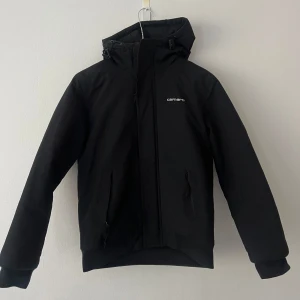 Svart jacka från Carhartt - Svart lätt fodrad vindjacka från Carhartt med rak passform, justerbar huva och dragkedja framtill. Jackan har två sidofickor med dragkedja och diskret Carhartt-logga på bröstet. Tillverkad i slitstarkt material som står emot vind och lätt regn. Säljer eftersom den är lite liten på mig, sparsamt använd och i fint skick.