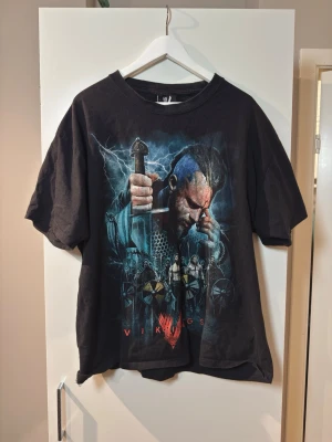 Svart Vikings t-shirt med tryck - (FITS AS XL) Cool svart t-shirt med stort tryck från serien Vikings på både fram- och baksidan. Framsidan har motiv av en viking med svärd och sköldar, medan baksidan visar en sköld och svärd. T-shirten har rund hals och korta ärmar. Perfekt för dig som gillar serien eller vill ha en unik statement-tee.
