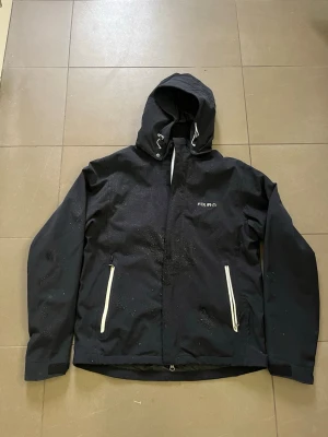 Svart regnjacka från Four: D - Snygg svart regnjacka från Four: D med huva och vita dragkedjor på fickorna. Jackan har vattenavvisande material och justerbar huva med snörning. Perfekt för regniga dagar och enkel att matcha med olika outfits.