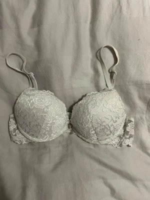 Vit spets-bh med dekorativ detalj - Säljer en vit bh med vadderade kupor och vacker spets över hela ytan. Bh:n har justerbara axelband och en liten dekorativ metallberlock framtill mellan kuporna.