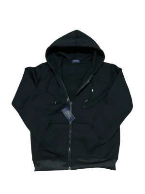 Svart hoodie från Polo Ralph Lauren - Svart hoodie med dragkedja från Polo Ralph Lauren. Klassisk design med huva och snören, två fickor fram och broderad logotyp på bröstet. Materialet är mjukt och bekvämt, perfekt för en avslappnad look.