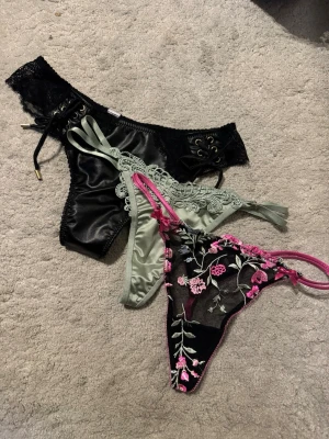 3 par stringtrosor i spets & satin – fina detaljer - Säljer ett set med tre olika stringtrosor i varierande modeller och material.  • Svarta satinstring med spetsdetaljer • Ljusgrön string med dekorativ spets • Svart string med rosa blommig brodyr och rosetter • Feminina detaljer och bekväm passform • Fint skick i storlek S/m  Perfekta om du vill uppdatera underklädeslådan med något snyggt och elegant ✨