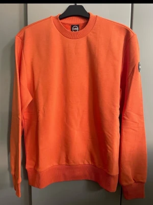 Orange sweatshirt från italienska Colmar  - Snygg orange sweatshirt från italienska Colmar med rund halsringning och ribbade muddar vid ärmslut och nederkant. Tröjan har en klassisk passform och en liten logotyp på ärmen. Perfekt för en avslappnad och trendig look. Storlek S herr men även tjejer kan ha 
