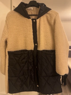 Beige teddyjacka med svarta detaljer - Teddyjacka i beige med stora svarta quiltade fickor och huva. Jackan har tryckknappar framtill och är gjord i mjukt, fluffigt material. Oversized passform och lång ärm, perfekt för kyliga dagar. Snygg kontrast mellan teddy och quiltad nylon.
