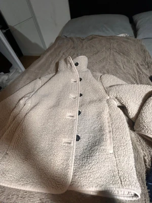 Beige teddyjacka med knappar - Mysig beige teddyjacka med hög krage och fem svarta knappar framtill. Jackan har två sidofickor och är gjord i fluffigt teddy-material som ger en avslappnad och trendig look. Perfekt för kalla dagar när du vill vara både varm och snygg.