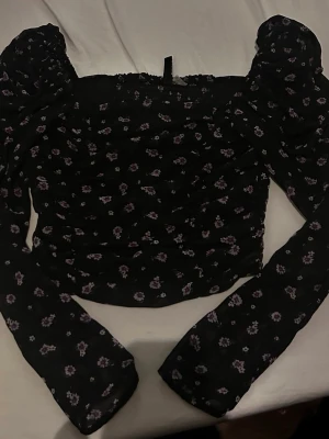 Svart blommig topp med puffärmar - Trendig svart topp med små lila blommor och långa puffärmar. Smockad över hela kroppen för en tight passform och fyrkantig urringning. Perfekt för dig som gillar romantisk och feminin stil. Materialet känns lätt och stretchigt.