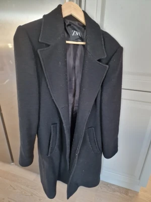 Svart kappa från Zara XL - Stilren svart kappa från Zara i storlek XL. Klassisk design med, två fickor framtill och slät yta. Perfekt för dig som vill ha en enkel men snygg look till hösten eller vintern. Lite trasig högr fickan, se bilden.