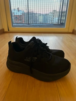 Svarta Skechers sneakers - Säljer ett par helsvarta Skechers sneakers med tjock sula och klassisk låg design. Skorna har snörning och är tillverkade i ett mesh-liknande material som andas. Perfekta för dig som gillar stilrena och bekväma skor till vardags.