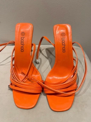Orange snörade pumps från Boohoo - Säljer ett par trendiga orange pumps från Boohoo med smala snören som lindas runt foten och höga stilettklackar. Skorna har fyrkantig tå och är tillverkade i ett glansigt syntetmaterial. Perfekta för dig som vill sticka ut med färg och cool design.
