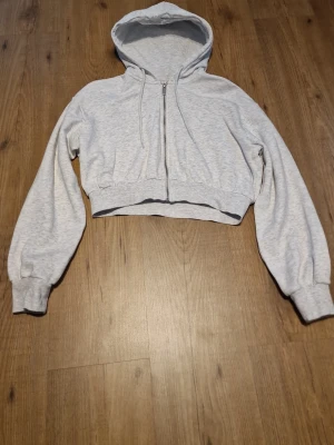 Ljusgrå cropped hoodie med dragkedja - Ljusgrå hoodie med huva och dragkedja framtill. Modellen är cropped. Perfekt för en avslappnad och trendig look. Materialet är mjukt och bekvämt, troligen bomullsblandning.