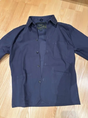 Mörkblå overshirt från Donce De La Sense - Stilren mörkblå overshirt med klassisk krage, tre utanpåliggande fickor och knäppning framtill. Tillverkad i ett kraftigare tyg som ger en schysst workwear-vibe. Perfekt att bära över en t-shirt eller hoodie. ALDRIG ANDVÄND. 