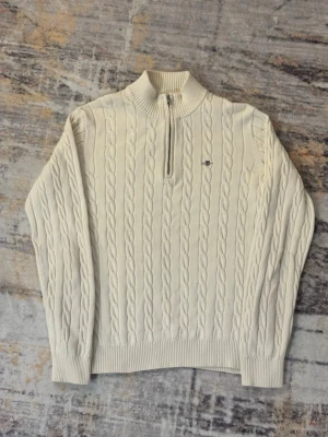 Gant halvzip - Vit GANT tröja med halvzip och ståkrage. Tröjan har ett diskret broderat märke på bröstet och ribbade muddar vid ärmslut och nederkant. Perfekt för lager-på-lager och en clean look.