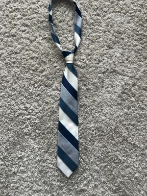 Randig slips i blått och vitt - Snygg slips med breda diagonala ränder i marinblått, ljusblått, grått och vitt. Tillverkad i ett glansigt material som känns lyxigt. Perfekt för att lyfta en skjorta med lite extra stil.
