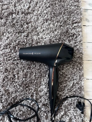 Remington Proline hårfön svart - Säljer en snygg och kraftfull hårfön från Remington Proline. Den är svart med eleganta gulddetaljer och har en klassisk form med ergonomiskt handtag. Perfekt för styling och torkning av håret. Elektrisk med sladd och stickkontakt. Sparsamt använd 