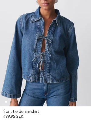 Ny Blå denimskjorta med snörning - Trendig blå denimskjorta med öppen front och snygga knytband. Skjortan har krage, vida ärmar och croppad passform. Perfekt för dig som gillar en avslappnad men cool stil. Helt ny och oanvänd 