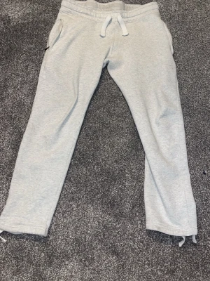 Ljusgrå russemerch byxor i storlek XS - Säljer ett par ljusgrå mjukisbyxor med vita dragsnören i midjan och fickor på sidorna. Byxorna har resår i midjan och mudd vid benslut samt en bakficka. Perfekta för chill dagar eller när du vill vara extra bekväm.