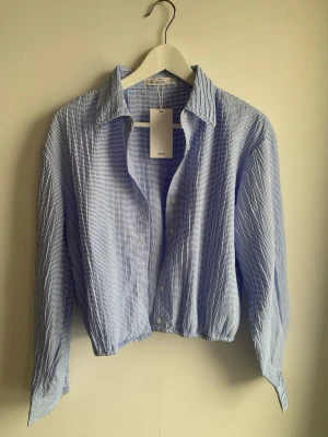 Blåvit croped skjorta från Mango L - Trendig blåvit cropped skjorta från Mango med tunna ränder och klassisk krage. Skjortan har långa ärmar med knappar vid ärmsluten och elastisk midja för en avslappnad look. Perfekt till jeans eller kjol för en cool och modern stil.
