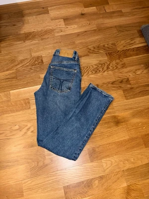 Tiger of Sweden  - Säljer ett par klassiska blå jeans med raka ben och hög midja. Jeansen har en bakficka och en läderpatch i midjan. Perfekta till en avslappnad stil och passar till det mesta. Materialet är kraftig denim i bomull.