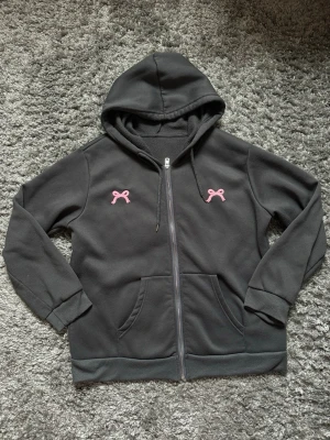 Mörkgrå hoodie med rosa rosetter - Säljer en mörkgrå hoodie med dragkedja och huva. På bröstet finns två broderade rosa rosetter som ger en söt touch. Hoodien har fickor framtill och ribbade muddar vid ärmar och nederkant. Perfekt för en avslappnad och trendig stil.