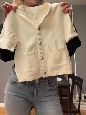 Vit kortärmad kofta med fickor - Säljer en söt vit kofta med korta ärmar och ribbade detaljer. Koftan har två framfickor och stora silvriga knappar framtill. Perfekt till jeans eller kjol för en clean och fräsch look.