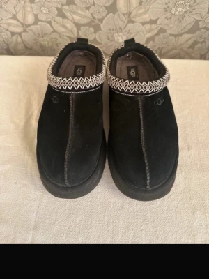 Svarta UGG tofflor i mocka - Säljer ett par svarta UGG tofflor i mjuk mocka med grått mönster runt öppningen. Insidan är fodrad med fluffigt material för extra komfort. Klassisk slip-on modell med diskret UGG-logga på sidan. Perfekta för att hålla fötterna varma hemma.