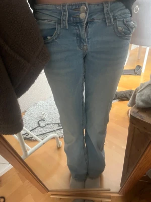 Ljusblå bootcut jeans med dubbla knappar - Säljer ett par ljusblå bootcut jeans med dubbla knappar i midjan och klassiska fem fickor. Jeansen har en lätt tvättad look och är tillverkade i denim med lite stretch för skön passform. Perfekta för en avslappnad och trendig stil.