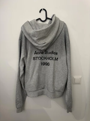 Grå hoodie från Acne Studios - Snygg grå hoodie från Acne Studios med tryck på ryggen där det står 'Acne Studios STOCKHOLM 1996'. Klassisk passform med stor huva och ribbade muddar. Perfekt för en avslappnad streetwear-look. Tillverkad i mjukt bomullsmaterial.