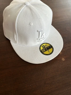 Vit LA 59FIFTY keps från New Era - Snygg vit snapback-keps från New Era, modell 59FIFTY, med LA-logga broderad framtill. Klassisk rak skärm och strukturerad passform. Tillverkad i bomullstyg och har den ikoniska guldstickern kvar på skärmen. Använd ca 2 gånger. Skicka gärna bud eller någon fråga om det behövs något mer tydligt!