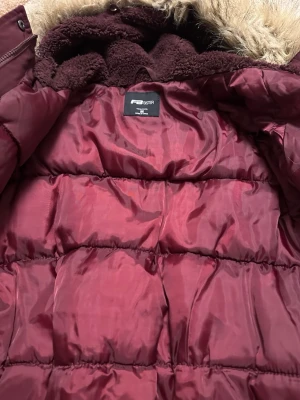 Vinröd pufferjacka från F&F - Snygg vinröd pufferjacka från F&F med mjukt foder och stor huva med fejkpäls. Jackan har quiltad design och dragkedja framtill. Perfekt för kalla vinterdagar och riktigt skön att bära. Materialet är glansigt och ger en modern look.